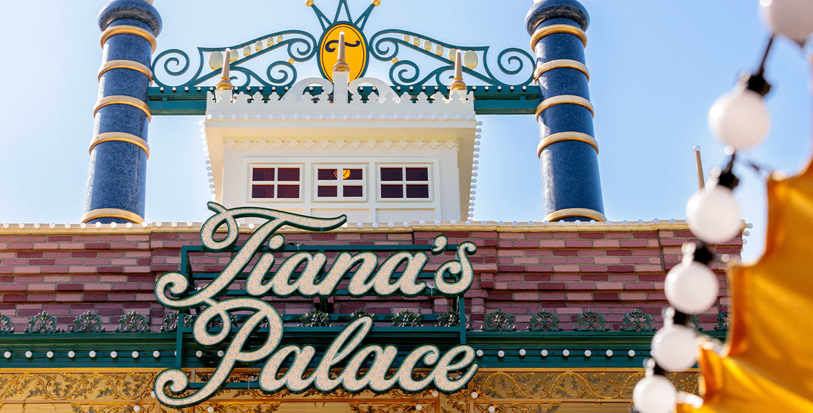 A close-up of the sign for Tiana’s Palace, written in cursive with unlit lights dotting the letters. The sign is attached to the top of the restaurant, in front of a red brick roof and blue smokestacks topped with crowns.