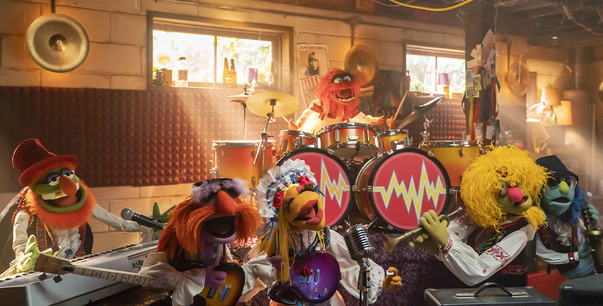 In an image from Disney+’s The Muppets Mayhem, The Electric Mayhem perform music excitedly, sunlight shining in beams through the windows behind them.