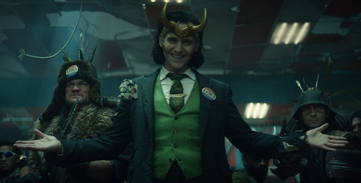 In an image from Disney+’s Loki, President Loki extends his arms with a confident expression while several other Loki variants stand behind him, preparing for battle.