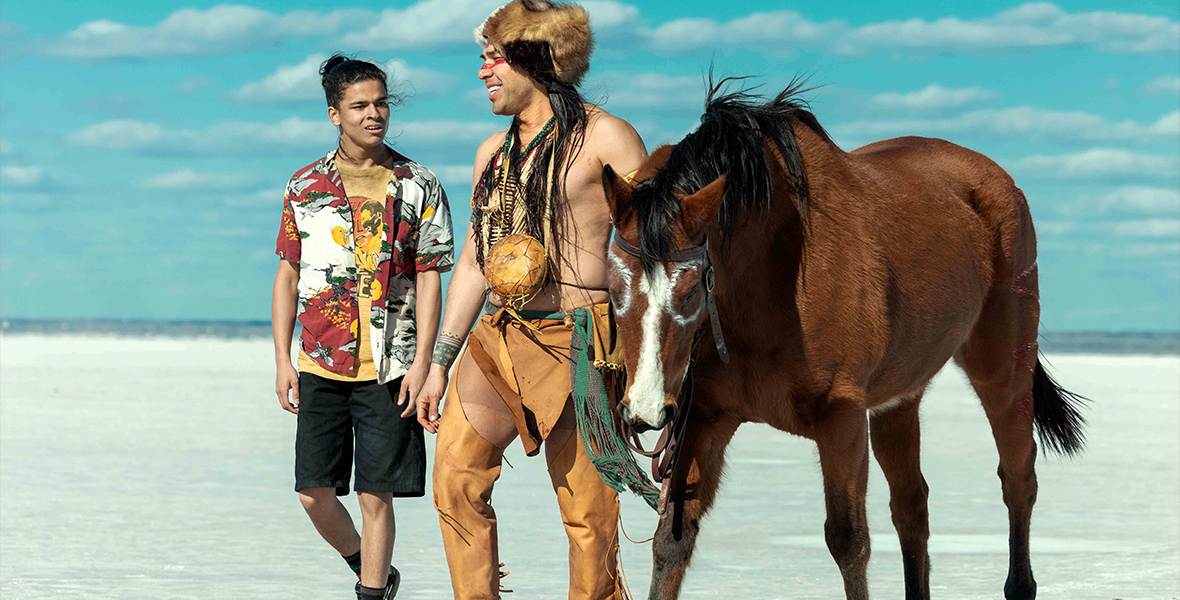 In an image from FX’s Reservation Dogs, Bear (D’Pharaoh Woon-A-Tai) and Spiritare (Dallas Goldtooth) are walking across a desert, in bright sunlight. Spiritare is wearing a traditional outfit, complete with fur cap, leather chaps, and beaded jewelry; he’s also leading a horse, who walks to his right. Bear is wearing a graphic button-up T-shirt and dark shorts. They’re talking as they walk.