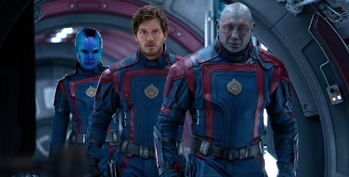 In an image from Marvel Studios’ Guardians of the Galaxy Vol. 3, from left to right, Nebula (Karen Gillan), Peter Quill/Star-Lord (Chris Pratt), and Drax (Dave Bautista) are walking along a corridor in their ship. They’re all wearing the traditional dark blue and red Guardians uniform, and have serious (or in the case of Peter, concerned) looks on their faces.