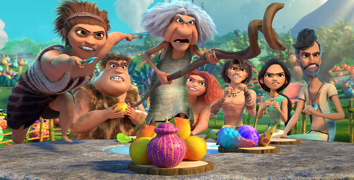 In an image from Hulu’s The Croods: Family Tree, the Crood family is seated or standing around a table. Several of them are angrily pointing at something off camera to the right. There are colorful fruits on the table, and behind them are rolling hills and unique vegetation.