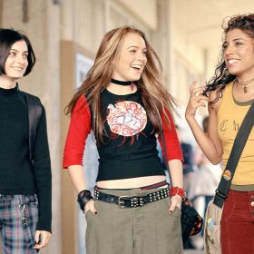 The characters from the film Freaky Friday stand side by side in a high school hallway including (from left to right), Anna who is played by Lindsay Lohan, Peg who is played by Haley Hudson, and Maddie who is played by Christina Vidal. Anna is wearing a black shirt with red ¾ length sleeves and a white, red, and purple circular patch on the front, baggy army green pants, a studded black belt, and a black choker. Peg is wearing a black turtleneck, red, blue, and white plaid pants, and a backpack with black straps. Maddie is wearing an army green tank top layered under a yellow tank top, maroon jeans, and a beige crossbody bag with a black strap.