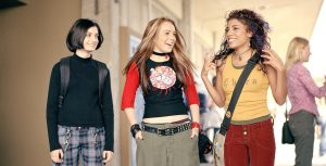 The characters from the film Freaky Friday stand side by side in a high school hallway including (from left to right), Anna who is played by Lindsay Lohan, Peg who is played by Haley Hudson, and Maddie who is played by Christina Vidal. Anna is wearing a black shirt with red ¾ length sleeves and a white, red, and purple circular patch on the front, baggy army green pants, a studded black belt, and a black choker. Peg is wearing a black turtleneck, red, blue, and white plaid pants, and a backpack with black straps. Maddie is wearing an army green tank top layered under a yellow tank top, maroon jeans, and a beige crossbody bag with a black strap.