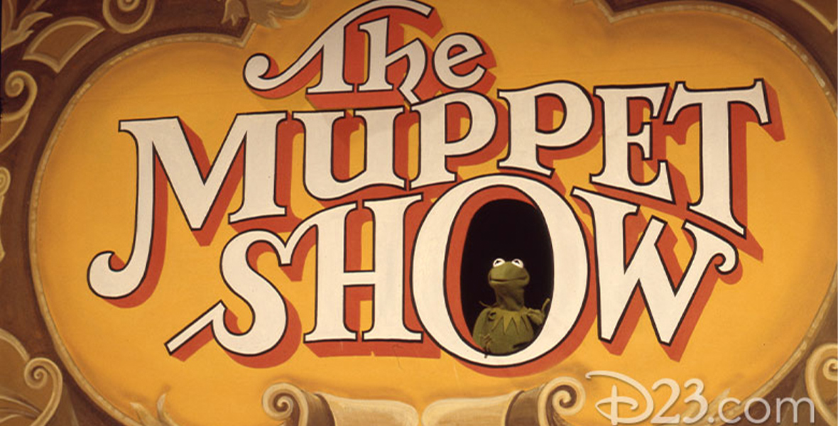 In an image from The Muppet Show, a stage drop with the logo of the show is seen, with a hole in the “O” of “Show,” in which Kermit the Frog can be seen. The logo itself is white lettering with a red shadow, on a yellow background, surrounded by golden leaves and decorative swirls.