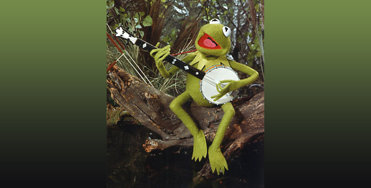 In an image from The Muppet Movie, Kermit the Frog is sitting on a log in a swamp, strumming a banjo and singing.