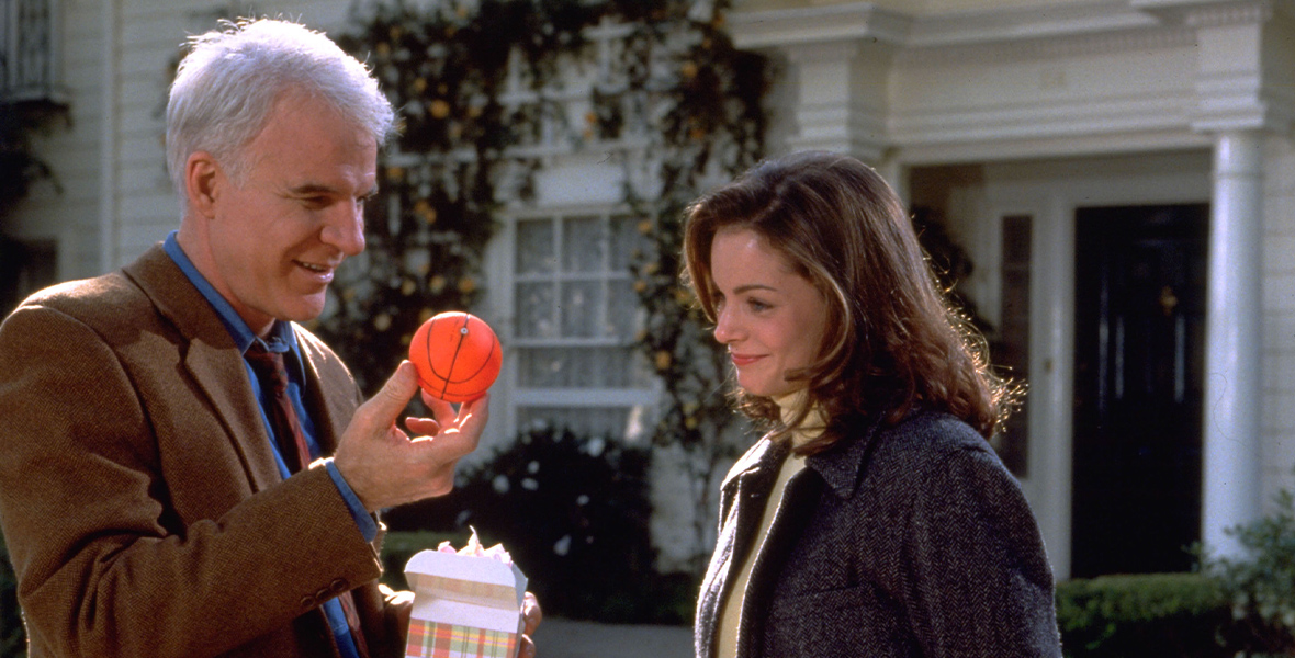 In an image from Father of the Bride Part II, George (Steve Martin) and Annie (Kimberly Williams-Paisley) are standing in front of the family home; it’s daytime, and it’s sunny outside. George is holding a box, out of which he has taken out a small basketball; in front of him is a baby carriage. They both are wearing blazers, and have wistful smiles on their faces.