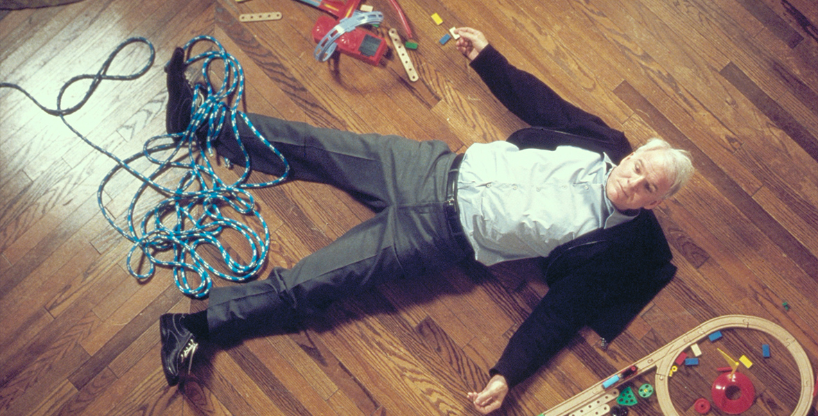 In an image from Cheaper by the Dozen, Tom Baker (Steve Martin) is lying on a wooden floor. He has fallen, and around him are various toys, as well as a long length of rope. He’s wearing dark pants, a white shirt, and a dark sweater. He looks like he’s in pain.