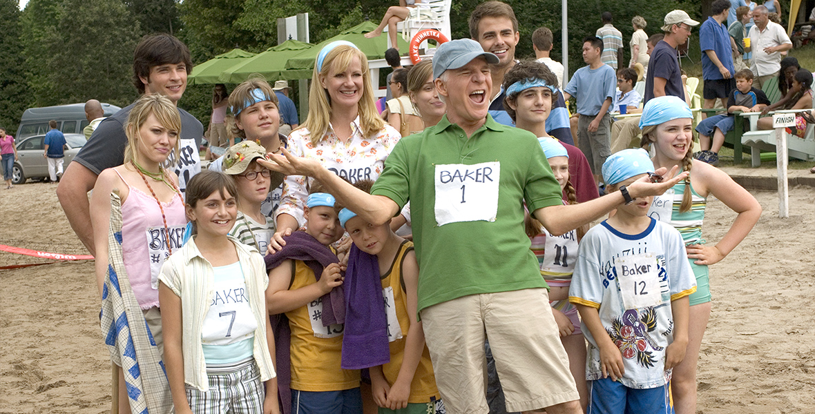 In an image from Cheaper by the Dozen 2, Tom Baker (Steve Martin) is surrounded by his large family, including wife Kate (Bonnie Hunt) and children such as Lorraine (Hilary Duff), Charlie (Tom Welling), Sarah (Alyson Stoner), and more. They’re standing on the sand, on the shore of a lake; other people are milling around behind them. They all have papers pinned to their shirts, with their names in large letters and numbers from 1 to 12. Tom has thrown his hands up as if he’s good-naturedly confronting someone offscreen.