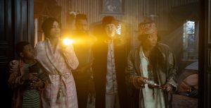 Travis (Chase Dillon), Gabbie (Rosario Dawson), Ben (LaKeith Stanfield), Father Kent (Owen Wilson), and Harriet (Tiffany Haddish) stand in the entrance to the “stretching room” in the film Haunted Mansion. All five are looking out at something offscreen, with Gabbie holding up a flashlight. Behind them, one of the iconic portraits from the Disney Parks Haunted Mansion attraction in its unstretched form is hanging on the wall.