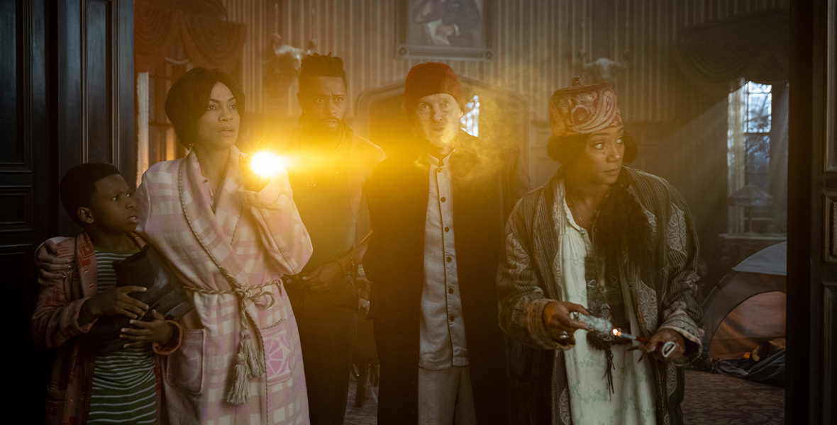Travis (Chase Dillon), Gabbie (Rosario Dawson), Ben (LaKeith Stanfield), Father Kent (Owen Wilson), and Harriet (Tiffany Haddish) stand in the entrance to the “stretching room” in the film Haunted Mansion. All five are looking out at something offscreen, with Gabbie holding up a flashlight. Behind them, one of the iconic portraits from the Disney Parks Haunted Mansion attraction in its unstretched form is hanging on the wall.