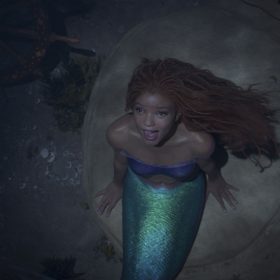 Ariel, played by Halle Bailey in the live-action The Little Mermaid, sits on a rock in the ocean and looks up while singing “Part of Your World” and wearing a purple, strapless top.