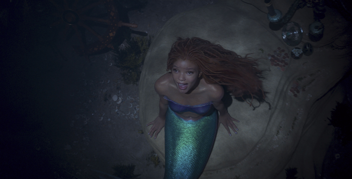Ariel, played by Halle Bailey in the live-action The Little Mermaid, sits on a rock in the ocean and looks up while singing “Part of Your World” and wearing a purple, strapless top.