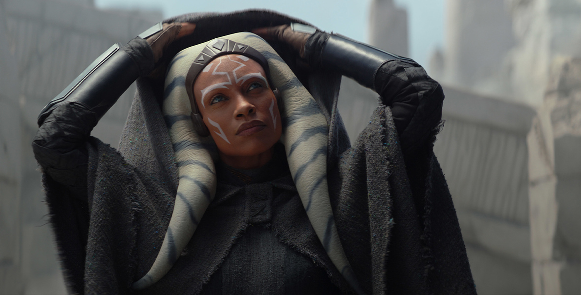 Ahsoka Tano (Rosario Dawson), from Star Wars: Ahsoka, removes the hood of her black cape. She is wearing a black shirt, black cape, and black leather lower arm braces as she looks to the side.