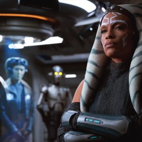Ahsoka Tano (Rosario Dawson), an orange-skinned Togrutan with white face markings, stands cross-armed in the hall of a spaceship. Behind her, slightly out of focus, is a blue-tinted hologram of the Twi’lek Hera Syndulla (Mary Elizabeth Winstead).