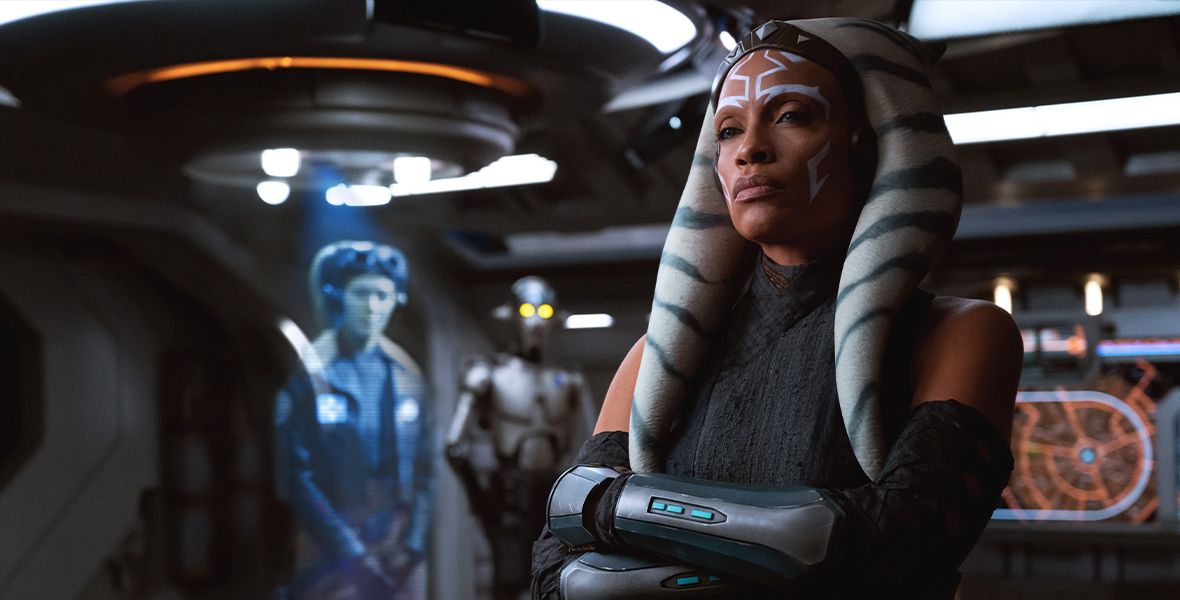 Ahsoka Tano (Rosario Dawson), an orange-skinned Togrutan with white face markings, stands cross-armed in the hall of a spaceship. Behind her, slightly out of focus, is a blue-tinted hologram of the Twi’lek Hera Syndulla (Mary Elizabeth Winstead).