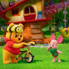 A still from Playdate with Winnie the Pooh, featuring Winnie the Pooh holding a yellow tricycle and wearing a bike helmet designed to look like a bee. Piglet stands next to him, wearing a green scarf and looking excitedly at the bike. They both stand in front of a whimsical treehouse.