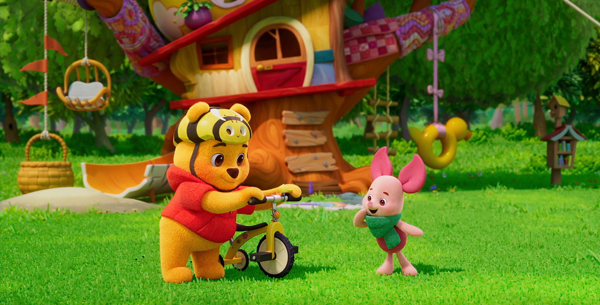 A still from Playdate with Winnie the Pooh, featuring Winnie the Pooh holding a yellow tricycle and wearing a bike helmet designed to look like a bee. Piglet stands next to him, wearing a green scarf and looking excitedly at the bike. They both stand in front of a whimsical treehouse.