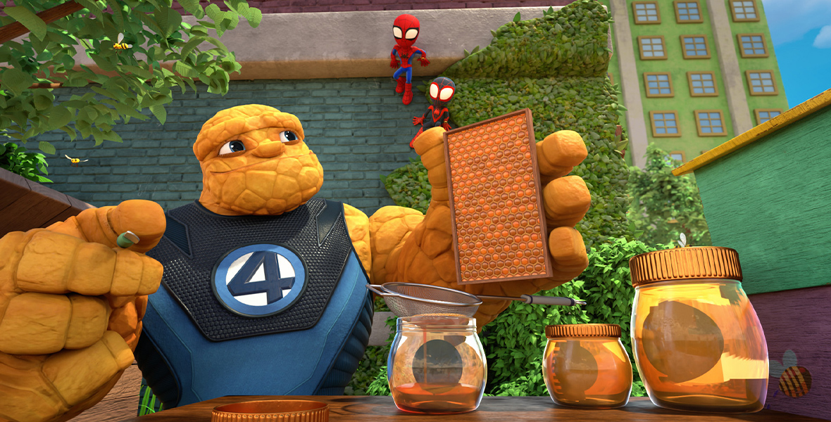 An image from the series Marvel’s Spidey and His Amazing Friends featuring The Thing sitting at a table and holding up honeycomb. On the table in front of him are jars of honey. Behind him, watching while stuck to the brick wall are Spidey (voiced by Benjamin Valic) and Miles Morales (voiced by Jakari Fraser), both in their costumes. 