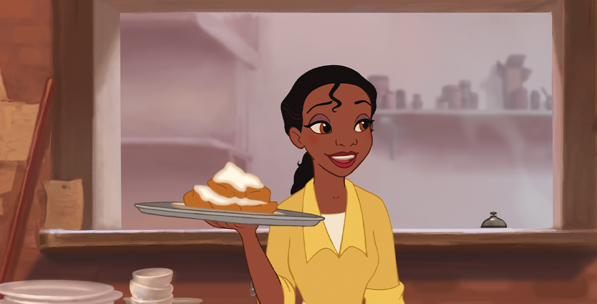 Tiana, from The Princess and the Frog, is standing in front of the window where food comes out in a restaurant. She is wearing a yellow-colored dress with a shirt underneath and her hair in a ponytail. She is holding a tray with a plate of biscuits and gravy upon it and looks to the side and smiles.