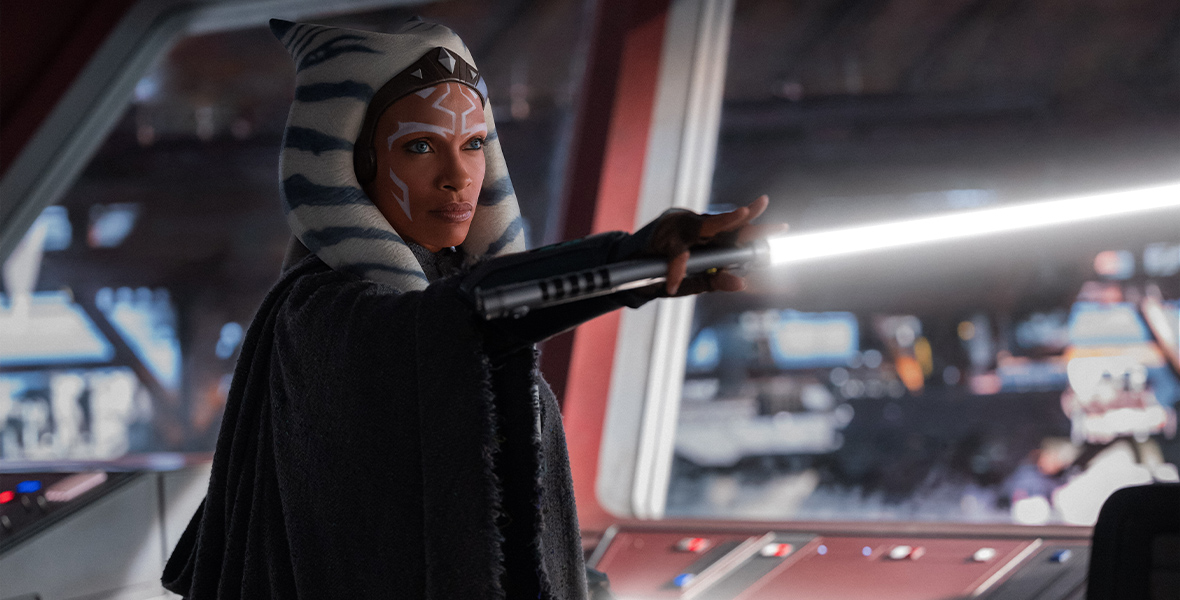In a scene from the second episode of Star Wars: Ahsoka, a robed Ahsoka Tano (Rosario Dawson) lifts a lightsaber that emits a white light. She is standing aboard a ship.