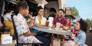 Two parents and their two children sit at a table while eating food outside a restaurant in Disney Springs. The son sits at the left of the table smiling while wearing blue pants and a collard shirt with Disney characters on it. He has a burger in front of him and a dark colored beverage in a glass. His mother is next to him and is smiling while wearing a yellow shirt under a white collared button down shirt that is open and blue jeans. The dad is next to her and is wearing a maroon short sleeve collard shirt with kaki shorts. He has a short glass in front of him with a large cube of ice and an orange beverage in it. The daughter sits at the right of the table while smiling at her brother across from her. She is wearing a white dress with read hearts on it, a light blue jean jacket, and Minnie Mouse ears. She has a stuffed animal of Minnie Mouse on her lap and a burger in front of her. In the middle of the table is a paper towel in a silver paper towel holder and in the background is a hanging sign that has “The Polite Pig” written on it.