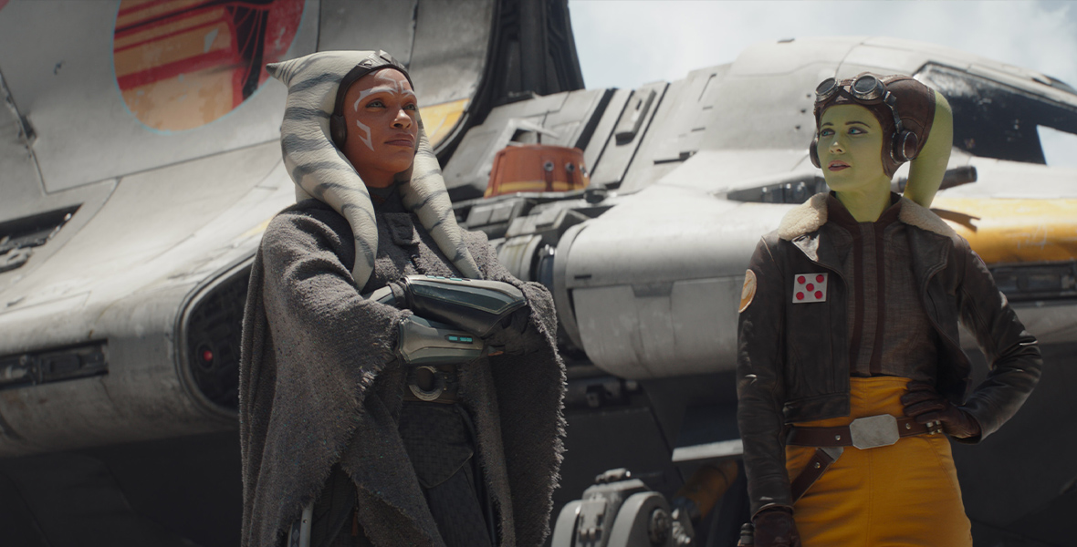 In an image from Star Wars: Ahsoka episode 2, from left to right, Ahsoka Tano (Rosario Dawson) and Hera Syndulla (Mary Elizabeth Winstead) are standing in front of a spacecraft, in which is seated Chopper the droid. Ahsoka’s arms are crossed and she’s looking at something off camera to the right; Hera is looking at Ahsoka. They are both wearing coats as if readying to travel somewhere.