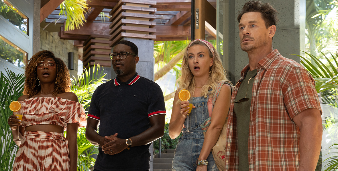In an image from Hulu’s Vacation Friends 2, from left to right, Emily (Yvonne Orji), Marcus (Lil Rey Howery), Kyla (Meredith Hagner), and Ron (John Cena) are standing in what appears to be the lobby of a tropical hotel. They’re all wearing beach/vacation wear, and have confused looks on their faces. Emily and Kyla are both holding drinks, and Kyla is also holding a removable baby car seat.