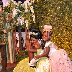 Prince Naveen smiles as Princess Tiana hugs two children wearing princess costumes.