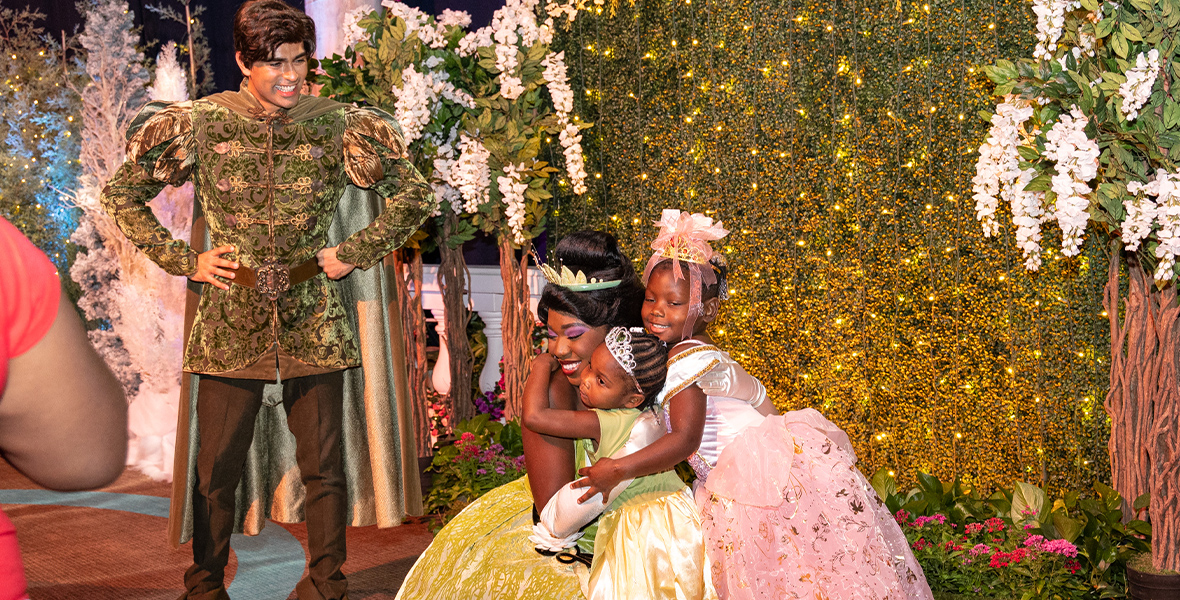 Prince Naveen smiles as Princess Tiana hugs two children wearing princess costumes.