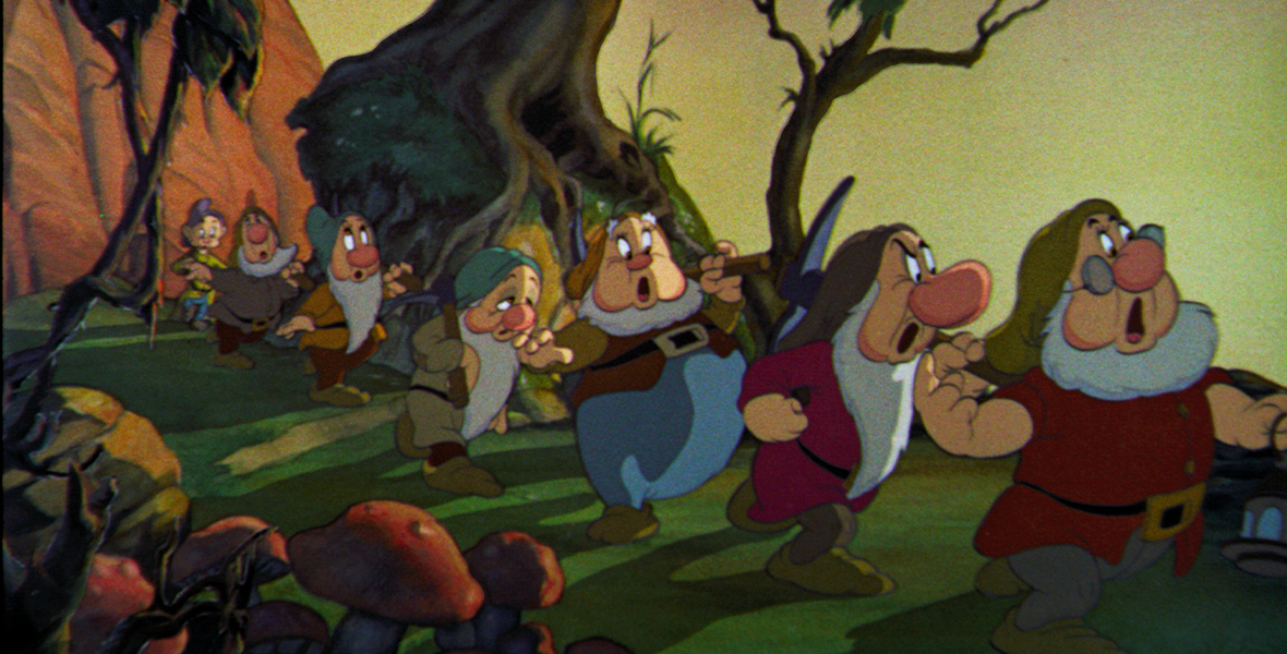 In an image from Walt Disney Animation Studios’ Snow White and the Seven Dwarfs, the dwarfs are seen marching along a hillside carrying their tools for mining. They’re in the middle of a song. A large tree can be seen behind them.