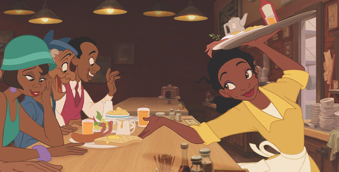 In an image from Walt Disney Animation Studios’ The Princess and the Frog, Tiana (voiced by Disney Legend Anika Noni Rose) is working at a restaurant and serving several customers seated at a counter to her left. She holds a tray aloft with her right hand, over her head, and is handing a plate to a customer with her left hand. She’s wearing a yellow dress and a white apron. Diner-type items can be seen on the counter, and the kitchen can be seen behind and to her right.