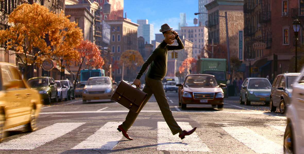In an image from Disney and Pixar’s Soul, Joe Gardner (voiced by Jamie Foxx) is walking briskly across a busy New York City street, within a striped crosswalk. He’s wearing a turtleneck, slacks, and a hat and is holding a briefcase in one hand and a phone up to his ear with the other. Cabs and other cars are whizzing by him on either side of the street.
