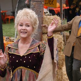 Aggie Cromwell from Halloween High, played by Debbie Reynolds, holds one hand in the air and one in front of her chest and smiles as she looks to the side while wearing a red, purple, and green collard dress with flowy sleeves and a maroon bodice that has gold stars and moons on it. Principal Flanigan, played by Clifton Davis, stands behind her looking surprised while wearing a blue collared shirt, beige blazer, yellow tie with red polka dots, and dark gray slacks. There are orange booths with hay stacks and skulls in the background.
