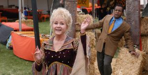 Aggie Cromwell from Halloween High, played by Debbie Reynolds, holds one hand in the air and one in front of her chest and smiles as she looks to the side while wearing a red, purple, and green collard dress with flowy sleeves and a maroon bodice that has gold stars and moons on it. Principal Flanigan, played by Clifton Davis, stands behind her looking surprised while wearing a blue collared shirt, beige blazer, yellow tie with red polka dots, and dark gray slacks. There are orange booths with hay stacks and skulls in the background.