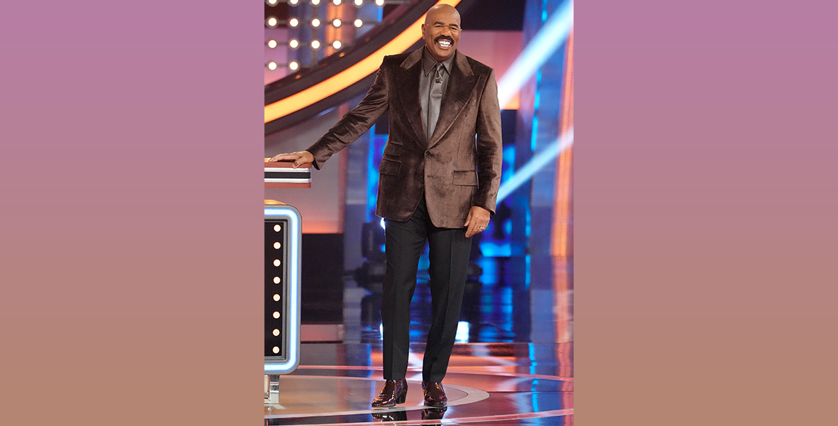 In an image from ABC’s Celebrity Family Feud, host Steve Harvey is standing on the brightly lit set, smiling. He’s wearing a brown velvet suit jacket and dark pants.