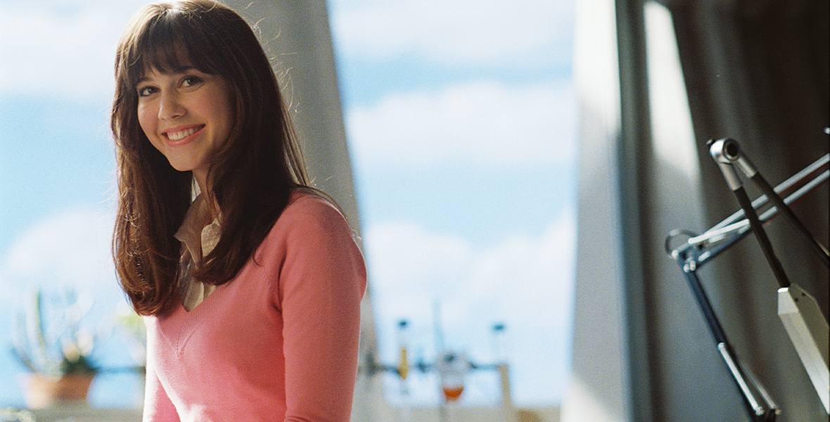 In an image from the film Sky High, Gwen Grayson (Mary Elizabeth Winstead) sits a desk in a Sky High classroom, wearing a pink sweater and smiling. Behind her is a window opening out onto the school campus.