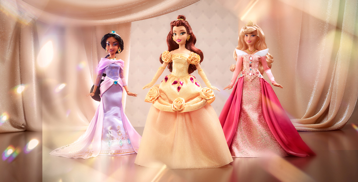 Fashion dolls of Jasmine, Belle, and Aurora against a rose gold curtain background. All three dolls are in bejeweled gowns.