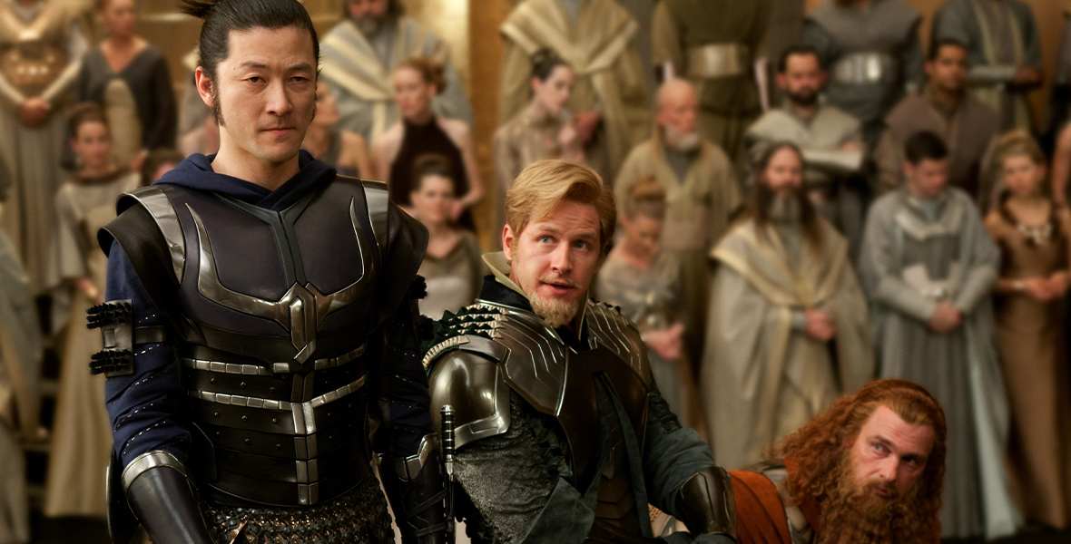 In an image from the film Thor, Hogun (Tadanobu Asano), Fandral (Josh Dallas), and Volstagg (Ray Stevenson) are all clad in armor, standing in front of a crowd inside the Asgardian palace. Hogun is standing, while Fandral and Volstagg are kneeling.