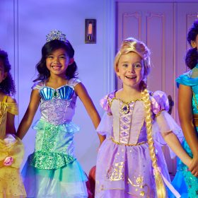 Four children stand in front of a house holding hands and dressed like Belle, Ariel, Rapunzel, and Jasmine.
