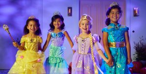 Four children stand in front of a house holding hands and dressed like Belle, Ariel, Rapunzel, and Jasmine.