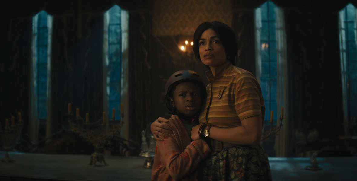 In an image from Disney’s Haunted Mansion, Travis (Chase Dillon) and Gabbie (Rosario Dawson) hug each other, looking at something off-camera in fear. They are standing in a dark, windowed hallway of a mansion.