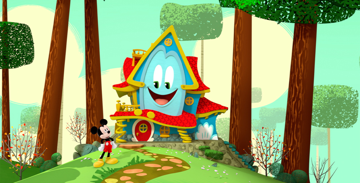 In a promotional image for Mickey Mouse Funhouse, the enchanted talking playhouse Funny (voice of Harvey Guillén) is seen in a forest setting, and Mickey Mouse (voice of Bret Iwan) is standing to their left, looking up at them. Trees surround Funny on either side, and there is a path leading up to Funny’s front door.