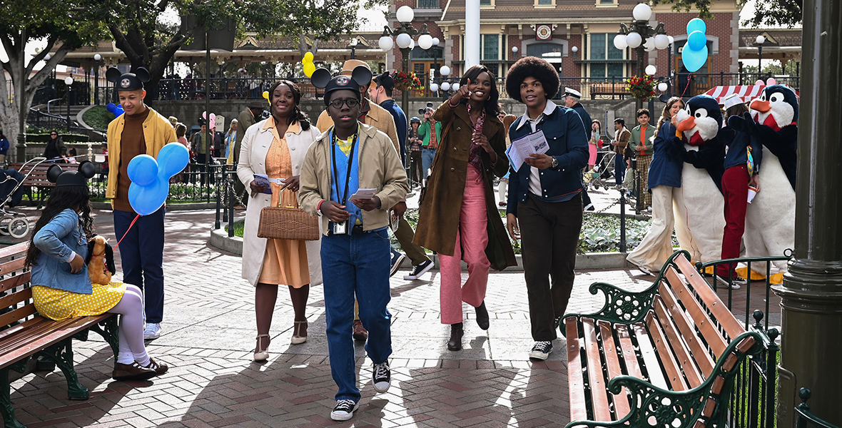 In an image from the second season finale episode of ABC’s The Wonder Years, Lillian (Saycon Sengbloh), Dean (Elisha “EJ” Williams), Kim (Laura Kariuki), and Bruce (Spence Moore II) are seen walking near the front of Disneyland Park, close to the Disneyland train station, which is seen in the background. Two costumed characters—penguins from Mary Poppins—are seen interacting with guests to the right. Benches are seen to the left and right, and other guests are milling about; some are holding Mickey ear balloons on strings. Dean is wearing Mickey ears.