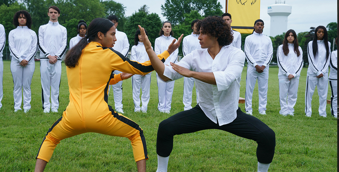 In an image from Hulu’s Miguel Wants to Fight, Miguel (Tyler Dean Flores), on the left, is dressed in a yellow jumpsuit a la Uma Thurman’s character in Kill Bill. He is readying to fight another person to the right, who’s dressed in a white button-up shirt and black pants. They are facing one another in fighting poses, with their arms up, on a grassy field. Behind them is a line of teenagers, all wearing white jumpsuits with black stripes down the arms and legs. A yellow flag with a line depiction of a lion is seen flying on a pole behind the teens to the right.