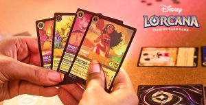 A person with fake nails featuring yellow french tips and red horizontal lines, is holding 4 playing cards in their hands. The front card has Moana on it with her name written under her pictures and a blurb titled We Can Fix It underneath. The card behind this card says Mikey on it and the rest of the card is hidden behind the Moana one. The two other cards are eligible as they are behind the Mikey card. On the table that the hands rest on is a card with Mickey on it and there is another card flipped over to reveal to the back of the card which is black, has a 4 point star in the middle of it with a circle around it, and pink and white swirls around the circle. There is a red, yellow, and green tint on the table.