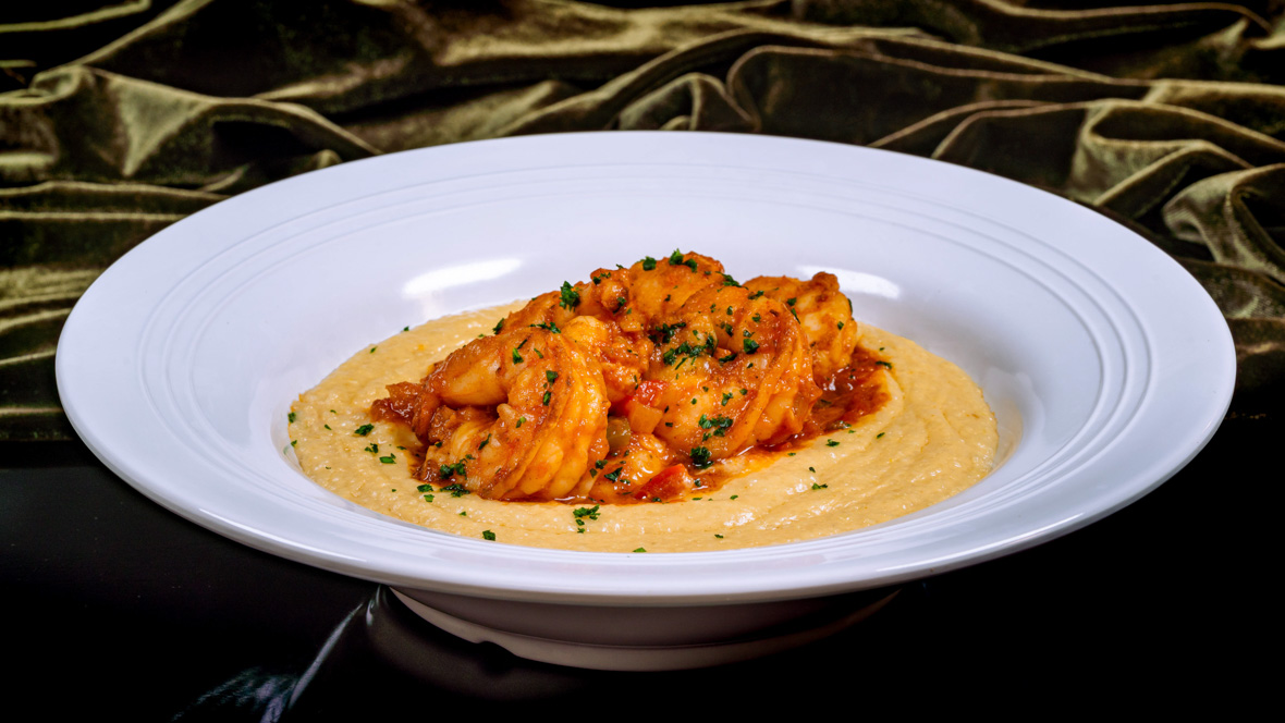 A white bowl holds a serving of Gulf Shrimp & Grits, as served at Tiana’s Palace. The reddish shrimp at the center are surrounded by cheesy yellow grits.