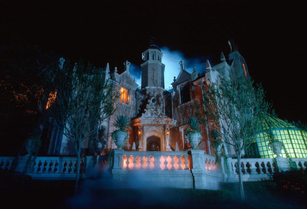 The exterior of Walt Disney World’s Haunted Mansion at nighttime. The mansion is covered in brick and designed in the Dutch Gothic style, surrounded by skinny tress. Yellow-orange light glows out of some of the windows.