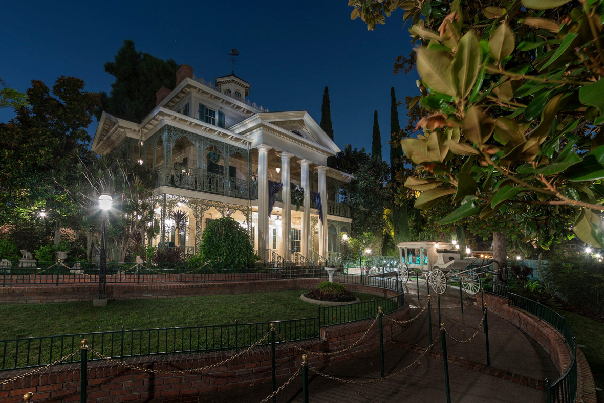 The exterior of Disneyland’s Haunted Mansion at nighttime. The mansion is white and features four tall columns in front, connecting the first two floors of the house. In front of the mansion is the attraction’s queue, a pet cemetery, and a horseless hearse.