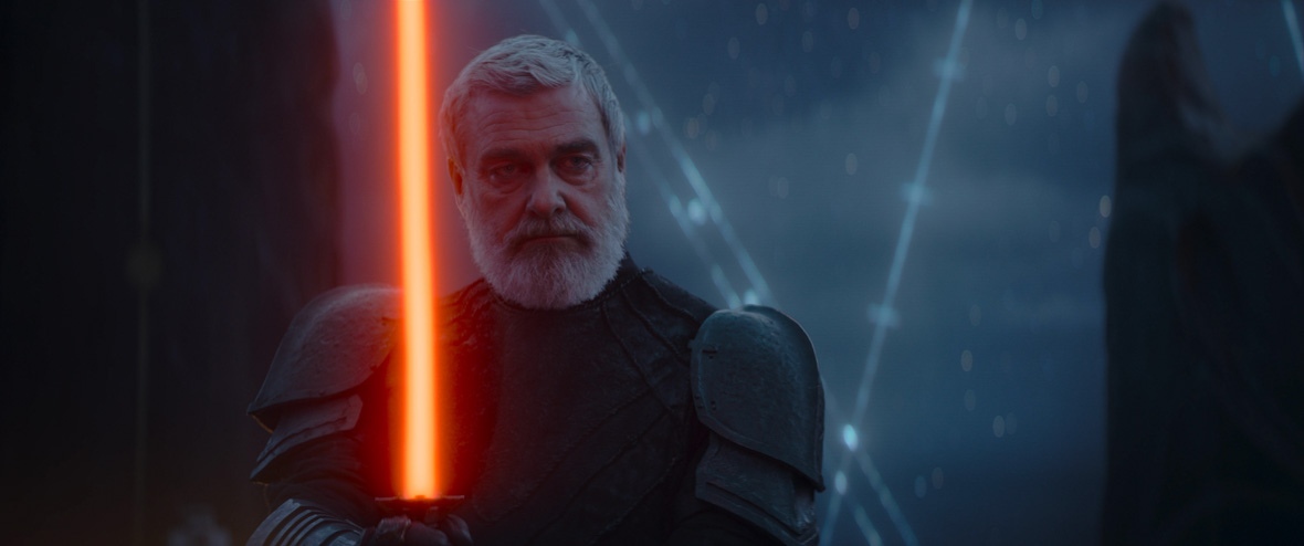 Baylan Skoll (Ray Stevenson), a pale human with grey hair and a beard, holds up his red lightsaber parallel to his face. He stands in partial darkness, his serious expression illuminated by the red glow of his lightsaber.